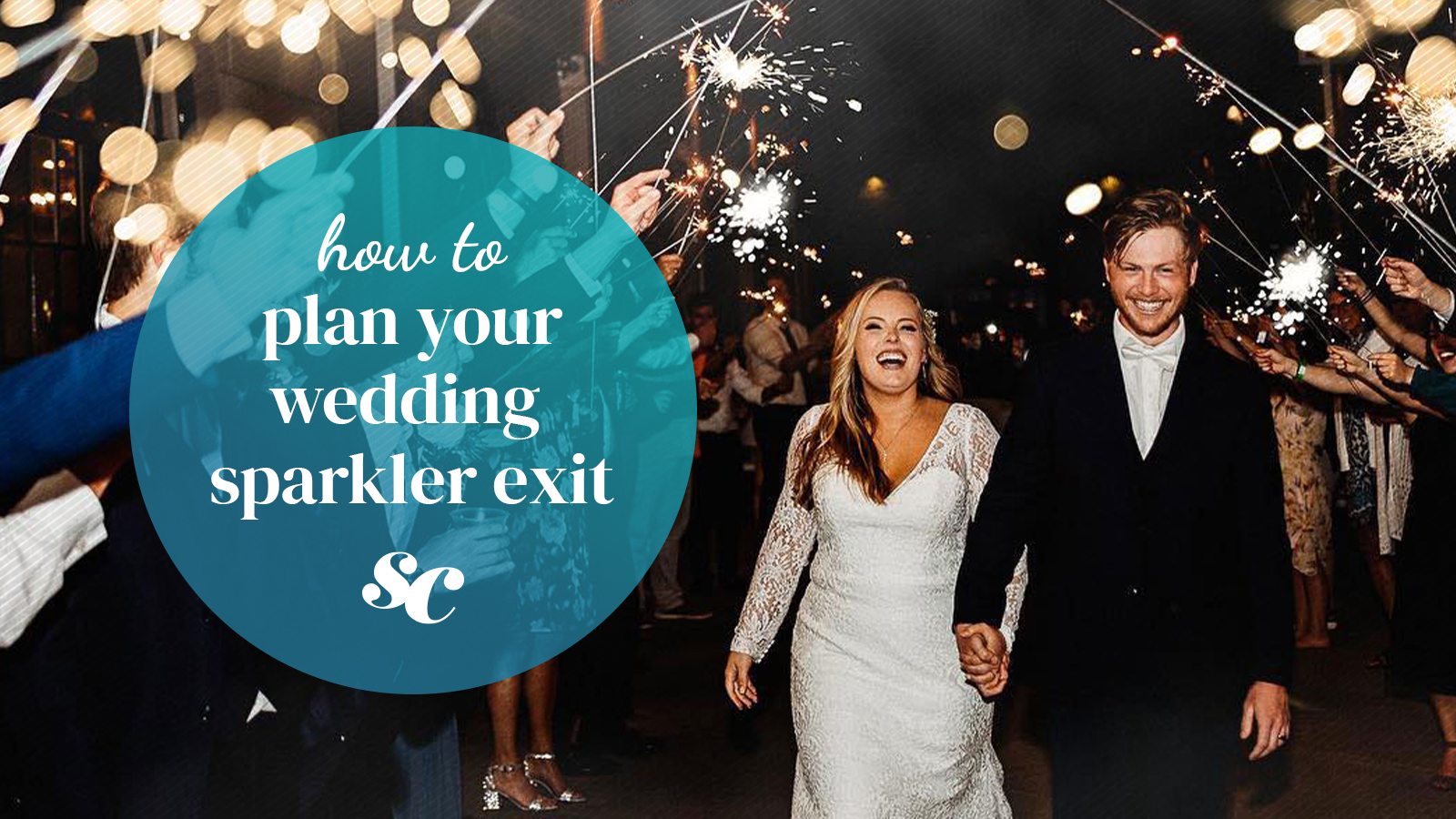 A smiling couple walks hand in hand between guests holding glowing sparklers.