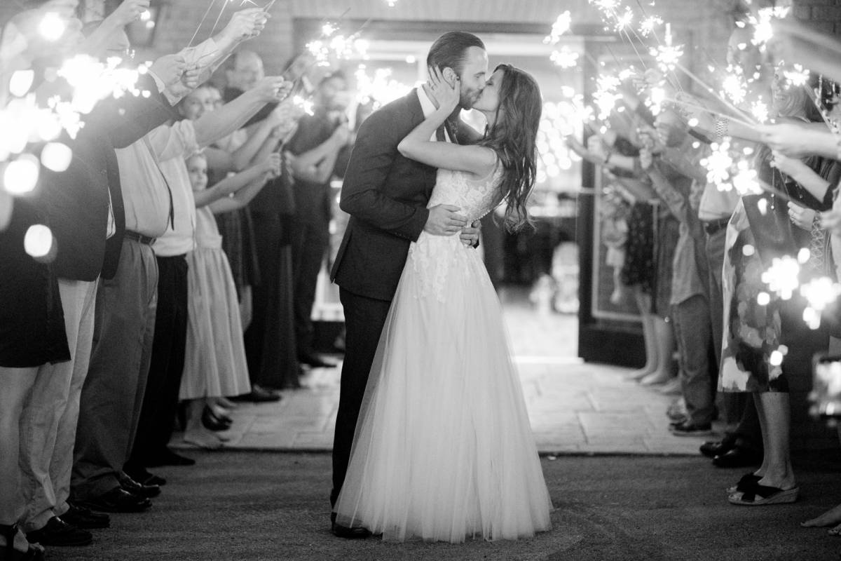 Black-and-white photograph of a couple pausing for a kiss between guests holding sparklers.